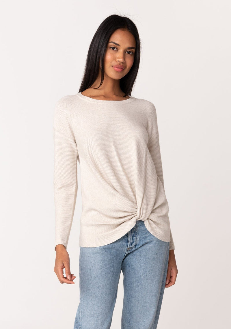 [Color: Heather Stone] A front facing image of a brunette model wearing a light heather stone pullover sweater in a ribbed textured knit. With long sleeves, a crew neckline, long sleeves, a relaxed fit, and a knot front waist detail. 