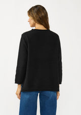 [Color: Black] A back facing image of a brunette model wearing a relaxed black honeycomb knit sweater with a round neckline, side vents and rolled edges.