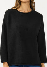 [Color: Black] A detail image of a brunette model wearing a relaxed black honeycomb knit sweater with a round neckline, side vents and rolled edges.