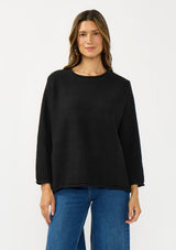 [Color: Black] A front facing image of a brunette model wearing a relaxed black honeycomb knit sweater with a round neckline, side vents and rolled edges.
