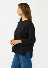 [Color: Black] A side facing image of a brunette model wearing a relaxed black honeycomb knit sweater with a round neckline, side vents and rolled edges.