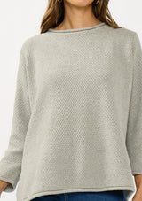 [Color: Cement] A detail image of a brunette model wearing a relaxed grey honeycomb knit sweater with a round neckline, side vents and rolled edges.