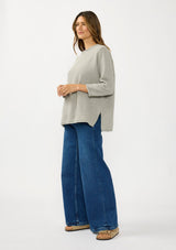 [Color: Cement] A side facing image of a brunette model wearing a relaxed grey honeycomb knit sweater with a round neckline, side vents and rolled edges.