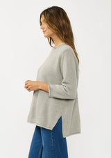 [Color: Cement] A side facing image of a brunette model wearing a relaxed grey honeycomb knit sweater with a round neckline, side vents and rolled edges.