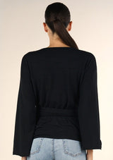 [Color: Black] A brunette model wearing a casual black wrap front top with long flowy sleeves, a v neckline, and an adjustable waist tie. 