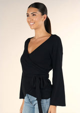 [Color: Black] A brunette model wearing a casual black wrap front top with long flowy sleeves, a v neckline, and an adjustable waist tie. 