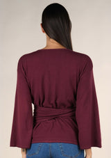 [Color: Burgundy] A brunette model wearing a casual burgundy wrap front top with long flowy sleeves, a v neckline, and an adjustable waist tie. 
