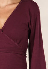 [Color: Burgundy] A brunette model wearing a casual burgundy wrap front top with long flowy sleeves, a v neckline, and an adjustable waist tie. 