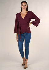 [Color: Burgundy] A brunette model wearing a casual burgundy wrap front top with long flowy sleeves, a v neckline, and an adjustable waist tie. 