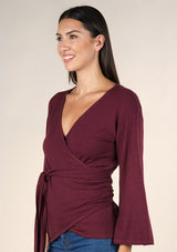[Color: Burgundy] A brunette model wearing a casual burgundy wrap front top with long flowy sleeves, a v neckline, and an adjustable waist tie. 