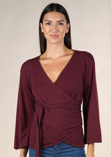 [Color: Burgundy] A brunette model wearing a casual burgundy wrap front top with long flowy sleeves, a v neckline, and an adjustable waist tie. 