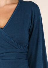[Color: Dark Sapphire] A detail image o of a brunette model wearing a casual blue wrap front top with long flowy sleeves, a v neckline, and an adjustable waist tie. 