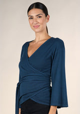 [Color: Dark Sapphire] A side facing image o of a brunette model wearing a casual blue wrap front top with long flowy sleeves, a v neckline, and an adjustable waist tie. 