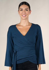 [Color: Dark Sapphire] A brunette model wearing a casual blue wrap front top with long flowy sleeves, a v neckline, and an adjustable waist tie. 