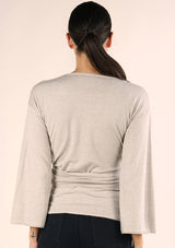 [Color: Heather Stone] A back facing image of a brunette model wearing a casual wrap front top with long flowy sleeves, a v neckline, and an adjustable waist tie. 