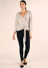 [Color: Heather Stone] A front facing image of a brunette model wearing a casual wrap front top with long flowy sleeves, a v neckline, and an adjustable waist tie. 