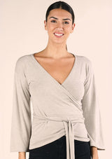 [Color: Heather Stone] A front facing image of a brunette model wearing a casual wrap front top with long flowy sleeves, a v neckline, and an adjustable waist tie. 