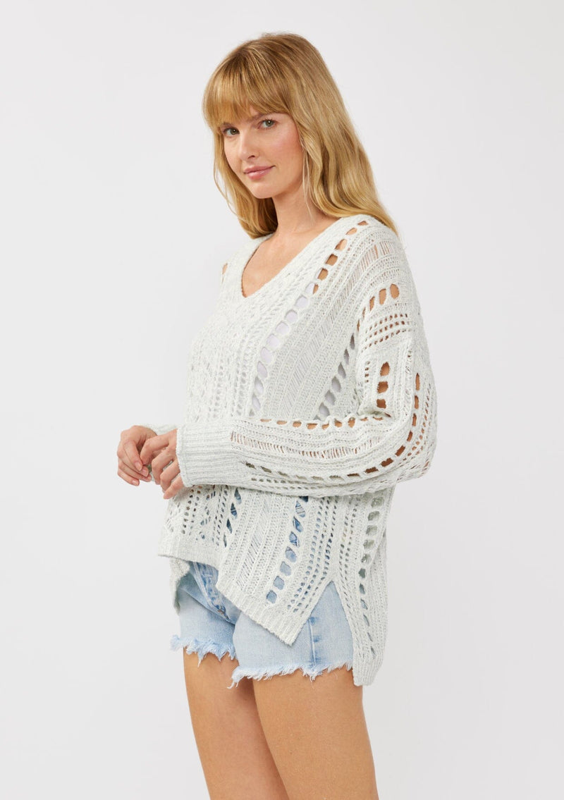 [Color: Blue] A side facing image of a blonde model wearing a light blue open knit crochet pullover sweater with a v-neckline long sleeves, dropped shoulders, and a side vents. Designed for a relaxed look and fit for the spring season. 