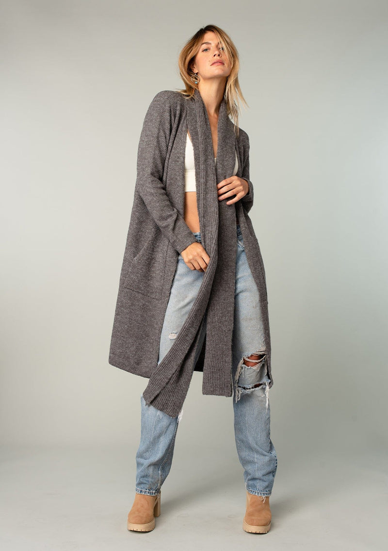 [Color: Olive] A model wearing a cozy dark grey long shawl cardigan. With long sleeves, side pockets, an open front, and attached scarf detail.