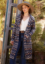 [Color: Navy/Camel] A front facing image of a brunette model standing outside wearing a sweater coat in a bohemian blue and brown chevron striped design. With long sleeves, a notched collar, a button front, side patch pockets, and a back vent.