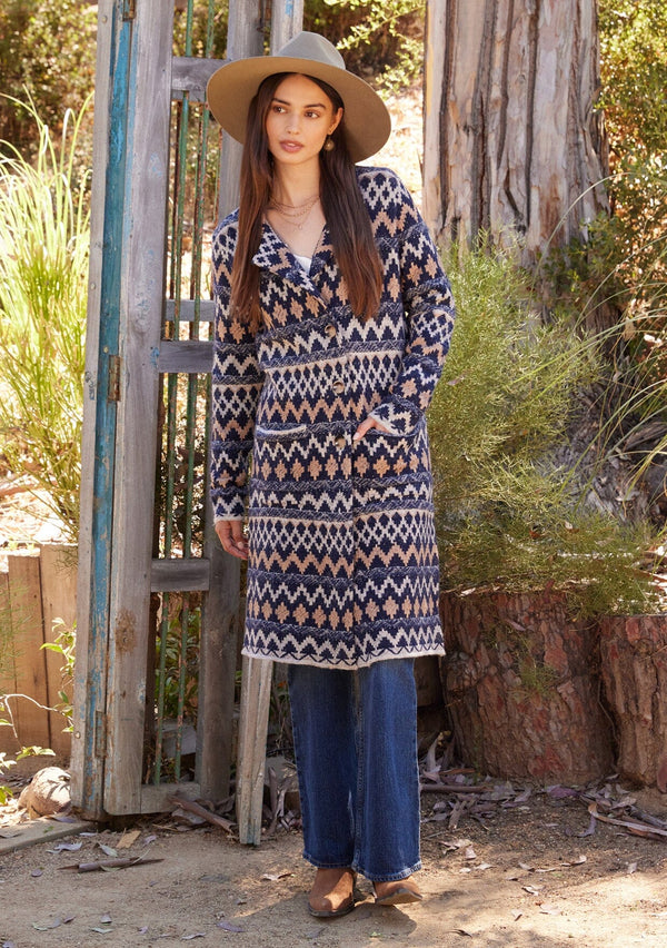 [Color: Navy/Camel] A full body front facing image of a brunette model standing outside wearing a sweater coat in a bohemian blue and brown chevron striped design. With long sleeves, a notched collar, a button front, side patch pockets, and a back vent.