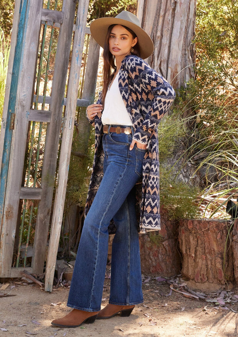 [Color: Navy/Camel] A full body side facing image of a brunette model standing outside wearing a sweater coat in a bohemian blue and brown chevron striped design. With long sleeves, a notched collar, a button front, side patch pockets, and a back vent.
