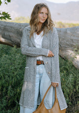 [Color: Taupe/Cream] A front facing image of a blonde model in the woods wearing a cozy and soft herringbone sweater coat with a notched lapel, button front, and patch pockets. 