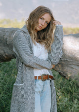 [Color: Taupe/Cream] A front facing image of a blonde model in the woods wearing a cozy and soft herringbone sweater coat with a notched lapel, button front, and patch pockets. 