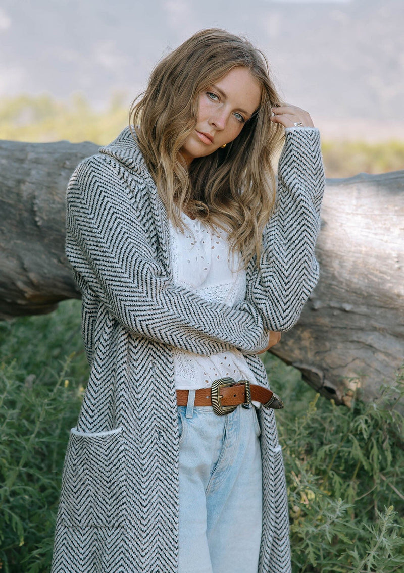 [Color: Taupe/Cream] A front facing image of a blonde model in the woods wearing a cozy and soft herringbone sweater coat with a notched lapel, button front, and patch pockets. 
