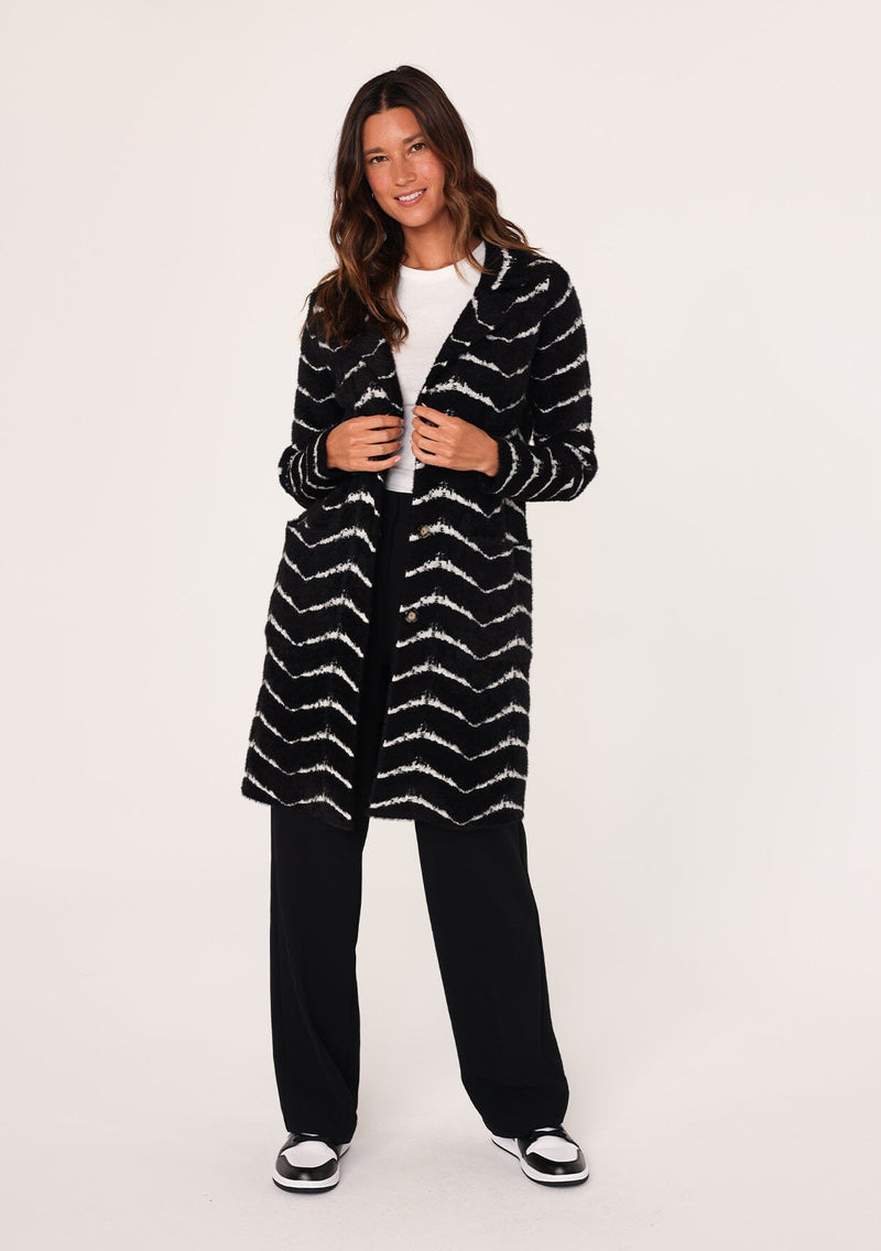[Color: Black/White] A full body front facing image of a brunette model wearing a soft and fuzzy sweater coat in a black and white chevron design. With a snap button front, side pockets, and a classic notched lapel.