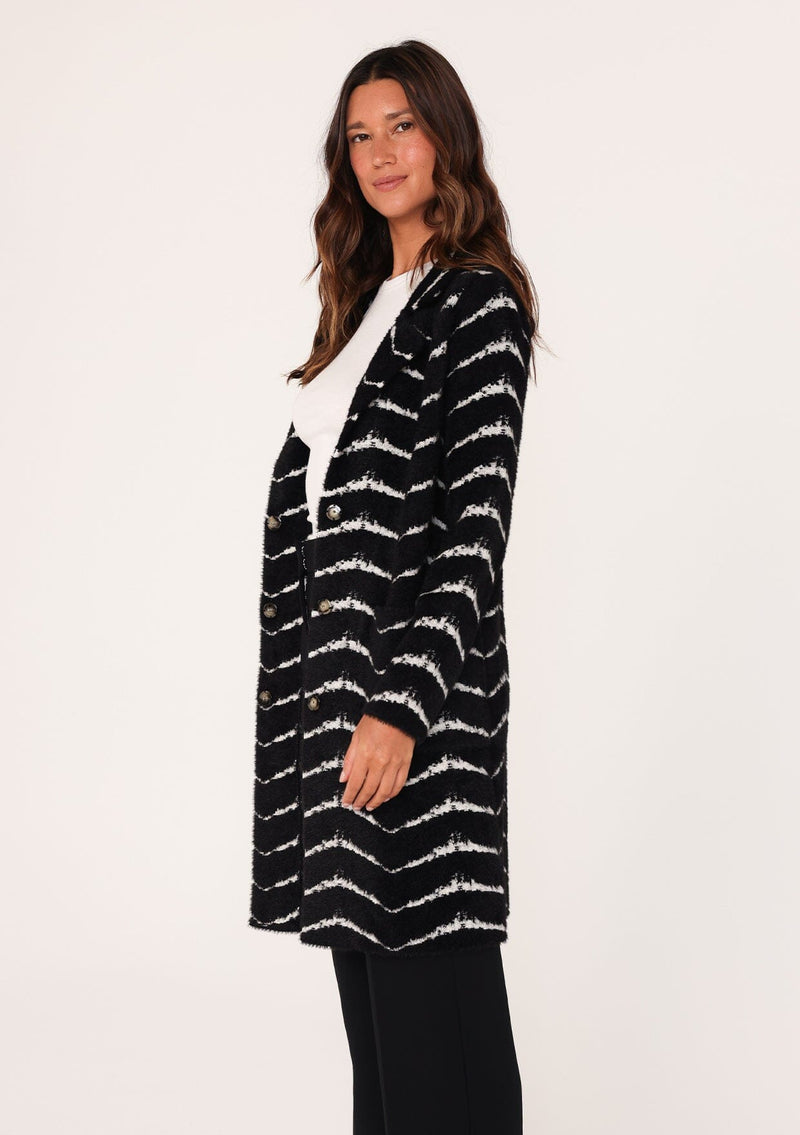 [Color: Black/White] A side facing image of a brunette model wearing a soft and fuzzy sweater coat in a black and white chevron design. With a snap button front, side pockets, and a classic notched lapel.