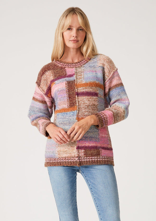 [Color: Pink Multi] A half body front facing image of a red headed model wearing a multi color pink patchwork knit sweater. With long sleeves, a crew neckline, and exposed seam details.