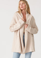 [Color: Heather Natural] A half body front facing image of a blonde model wearing an oversized sweater coat in ivory. With an oversized hood, long sleeves, an open front, side pockets, and a mid length. 
