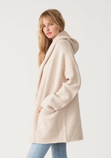 [Color: Heather Natural] A side facing image of a blonde model wearing an oversized sweater coat in ivory. With an oversized hood, long sleeves, an open front, side pockets, and a mid length. 