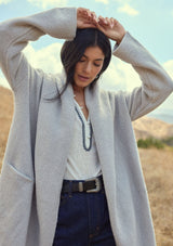 [Color: Heather Grey] A front facing image of a brunette model outside wearing an oversized sweater coat in heather grey. With an oversized hood, long sleeves, an open front, side pockets, and a mid length.   A minimalist coatigan styled for the fall and winter season.