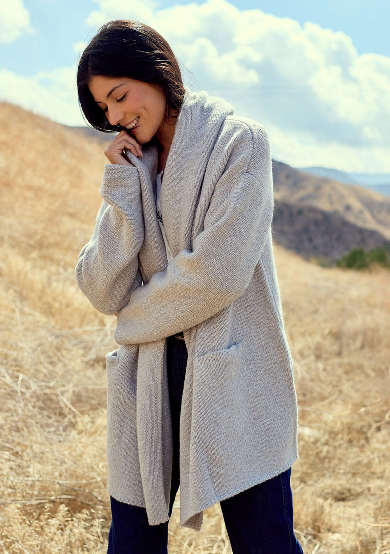 [Color: Heather Grey] A front facing image of a brunette model outside wearing an oversized sweater coat in heather grey. With an oversized hood, long sleeves, an open front, side pockets, and a mid length.   A minimalist coatigan styled for the fall and winter season.