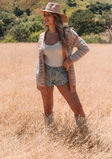 [Color: Mocha/Natural] A front facing image of a blonde model standing in a field wearing a bohemian fall cardigan in a speckled loop knit stripe. With long sleeves, side pockets, an open front, and contrast brown ribbed trim.