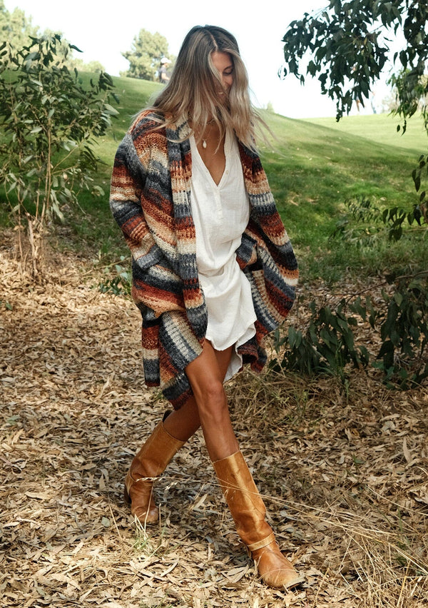 [Color: Rust/Navy] A front facing image of a blonde model wearing a bohemian blue and rust striped mid length cardigan. With long sleeves, an open front, side pockets, and an oversize hood.