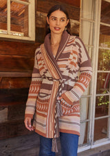 [Color: Taupe/Blush] A full body front facing image of a brunette model standing outside wearing a cozy fall bohemian cardigan in a pink and brown southwestern style pattern. With long sleeves, a shawl collar, side pockets, and a braided waist belt.