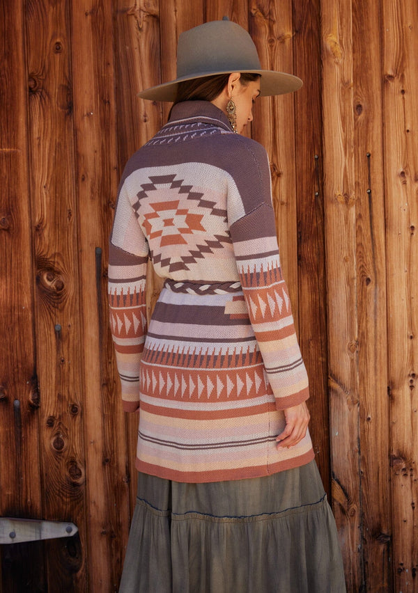 [Color: Taupe/Blush] A back facing image of a brunette model standing outside wearing a cozy fall bohemian cardigan in a pink and brown southwestern style pattern. With long sleeves, a shawl collar, side pockets, and a braided waist belt.