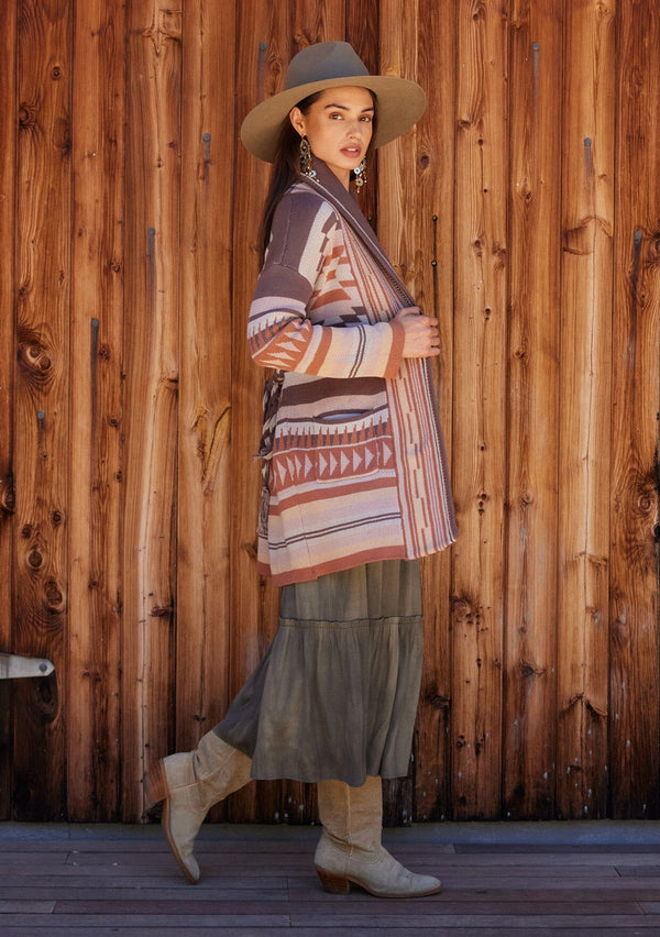 [Color: Taupe/Blush] A full body front facing image of a brunette model standing outside wearing a cozy fall bohemian cardigan in a pink and brown southwestern style pattern. With long sleeves, a shawl collar, side pockets, and a braided waist belt.