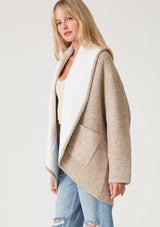 [Color: Natural] A side facing image of a blonde model wearing an oversize brown and ivory two tone knit cardigan. With an oversized shawl collar, an open front, and side pockets. 