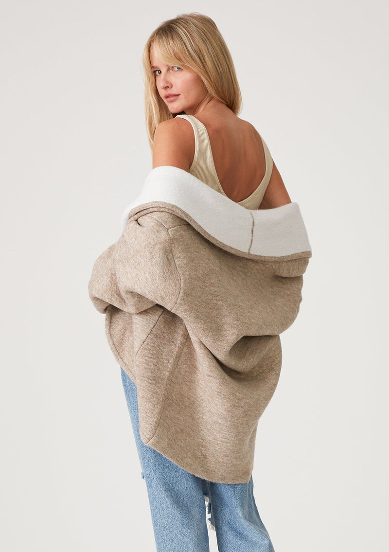 [Color: Natural] A half body back facing image of a blonde model wearing an oversize brown and ivory two tone knit cardigan. With an oversized shawl collar, an open front, and side pockets. 