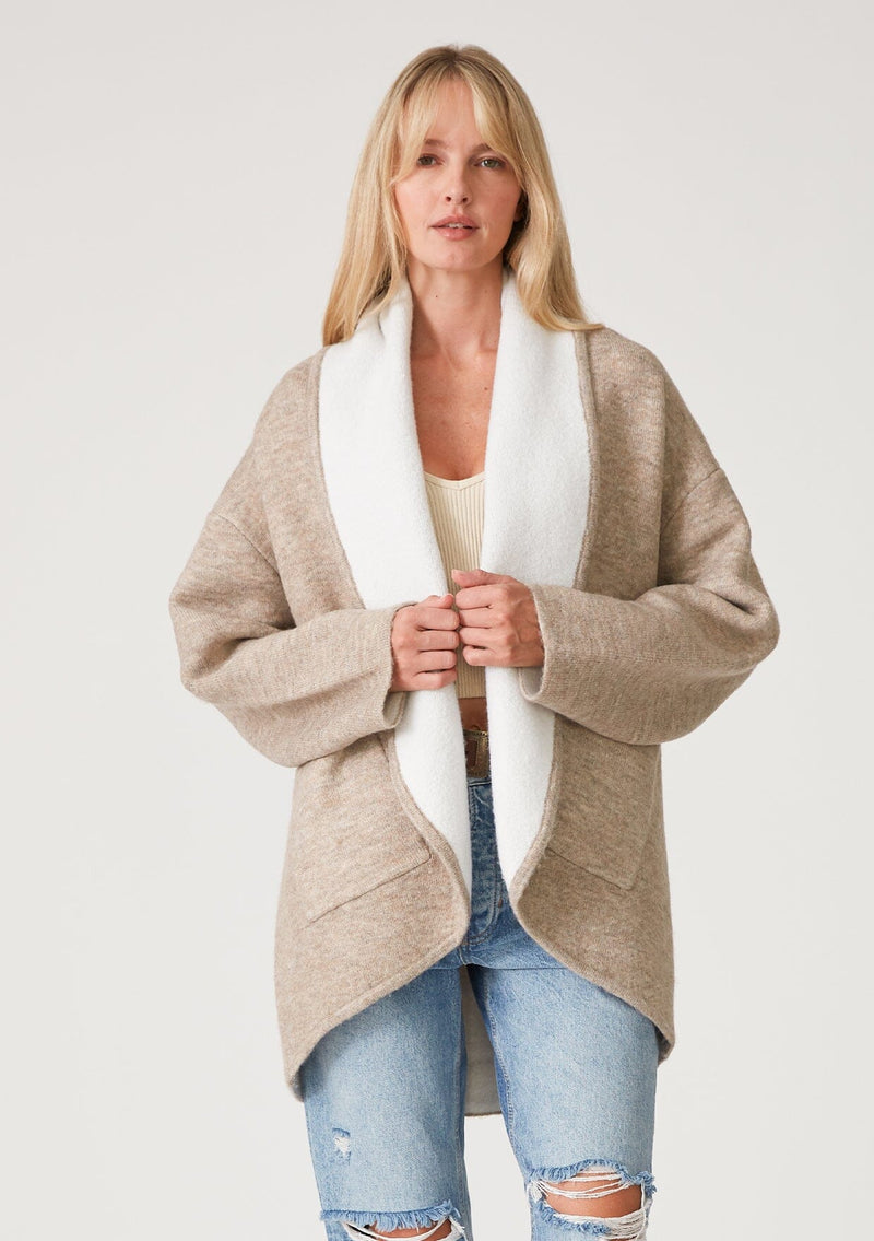 [Color: Natural] A front facing image of a blonde model wearing an oversize brown and ivory two tone knit cardigan. With an oversized shawl collar, an open front, and side pockets. 