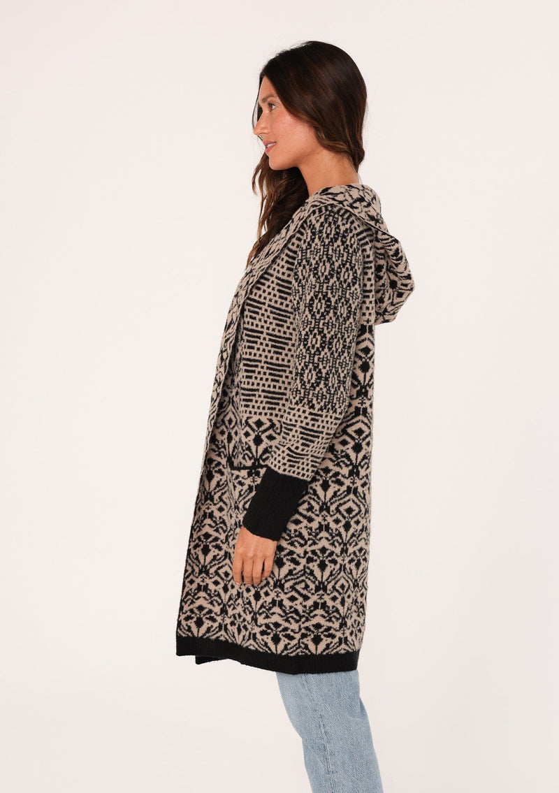 [Color: Taupe/Black] A side facing image of a brunette model wearing a soft boho cardigan in a brown and black abstract pattern. With a hoodie, an open front, a mid length hemline, and side pockets. 