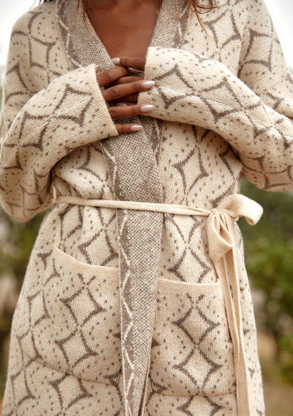 [Color: Natural/Taupe] A close up front facing image of a brunette model wearing a soft mid length cardigan sweater coat in an ivory diamond jacquard. With long sleeves, side pockets, and a belted tie waist.
