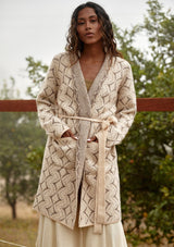 [Color: Natural/Taupe] A half body front facing image of a brunette model wearing a soft mid length cardigan sweater coat in an ivory diamond jacquard. With long sleeves, side pockets, and a belted tie waist.