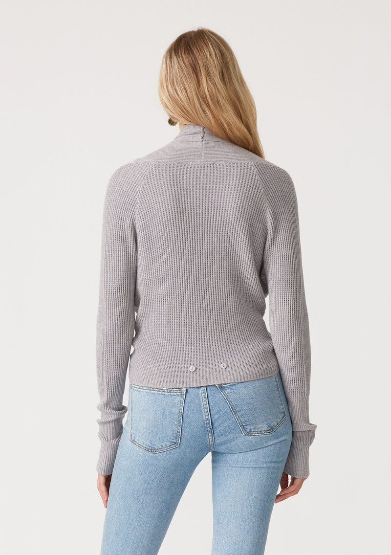 [Color: Heather Grey] A back facing image of a blonde model wearing a heather grey waffle knit wrap sweater with long sleeves, a v neckline, and a button closure at the back. The long ties can be styled in multiple ways. 
