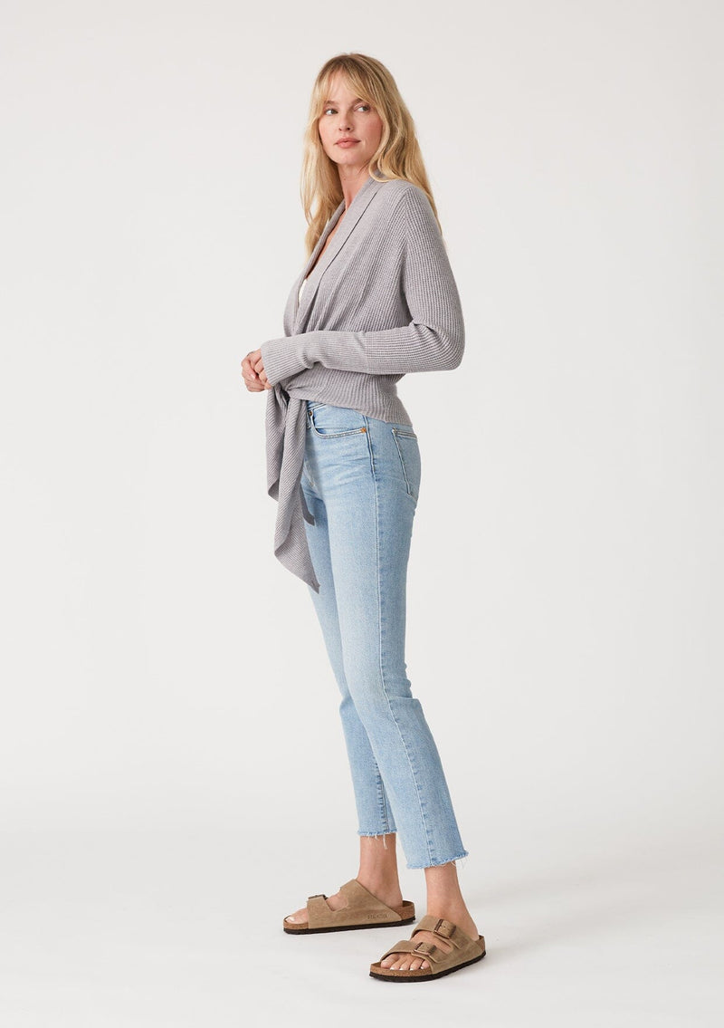 [Color: Heather Grey] A side facing image of a blonde model wearing a heather grey waffle knit wrap sweater with long sleeves, a v neckline, and a button closure at the back. The long ties can be styled in multiple ways. 