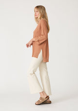 [Color: Clay] A side facing image of a blonde model wearing a clay brown lightweight sweater tunic top. With three quarter length sleeves, a button roll tab, a v neckline, exposed seam details, a long tunic length, and side vents. 